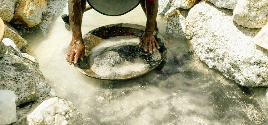 ชายยืนอยู่ในลำธารถือถังน้ำ สายน้ำไหลผ่านและมีต้นไม้เขียวขจีอยู่รอบข้าง