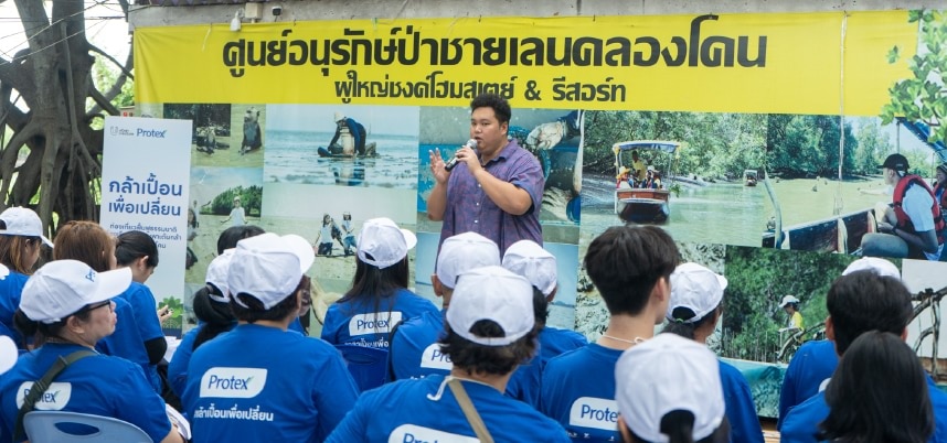 ชายสวมเสื้อเชิ้ตสีน้ำเงินกำลังกล่าวสุนทรพจน์ต่อกลุ่มผู้ฟังที่นั่งอยู่ด้านหน้าเขา