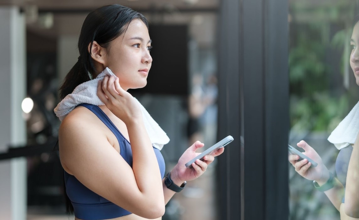 ผู้หญิงออกกำลังกายเช็ดเหงื่อด้วยผ้าและถือสมาร์ทโฟนในฟิตเนส