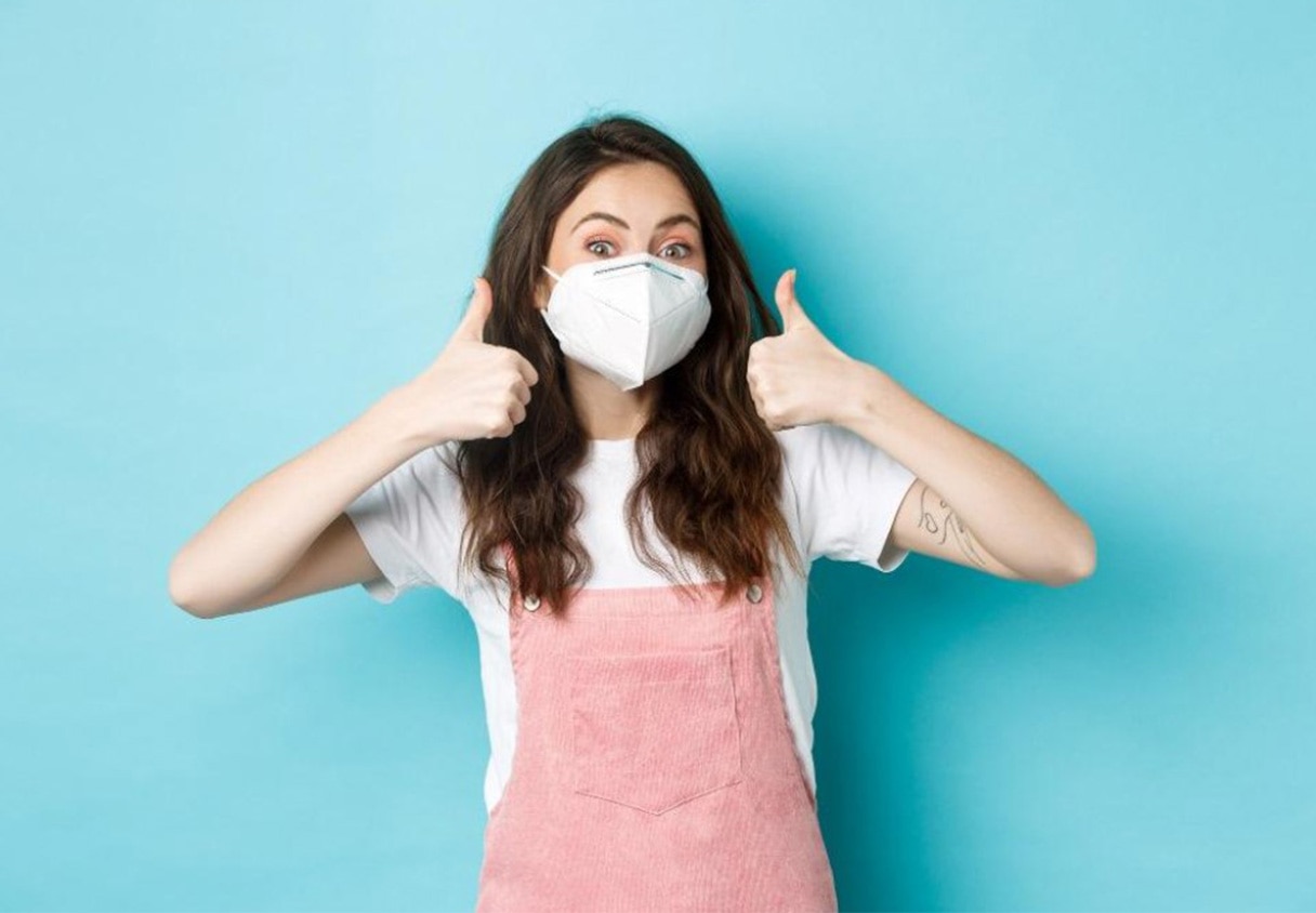 หญิงสาวสวมหน้ากากอนามัยสีขาว ยิ้มและทำท่าทางชูนิ้วโป้งทั้งสองข้าง