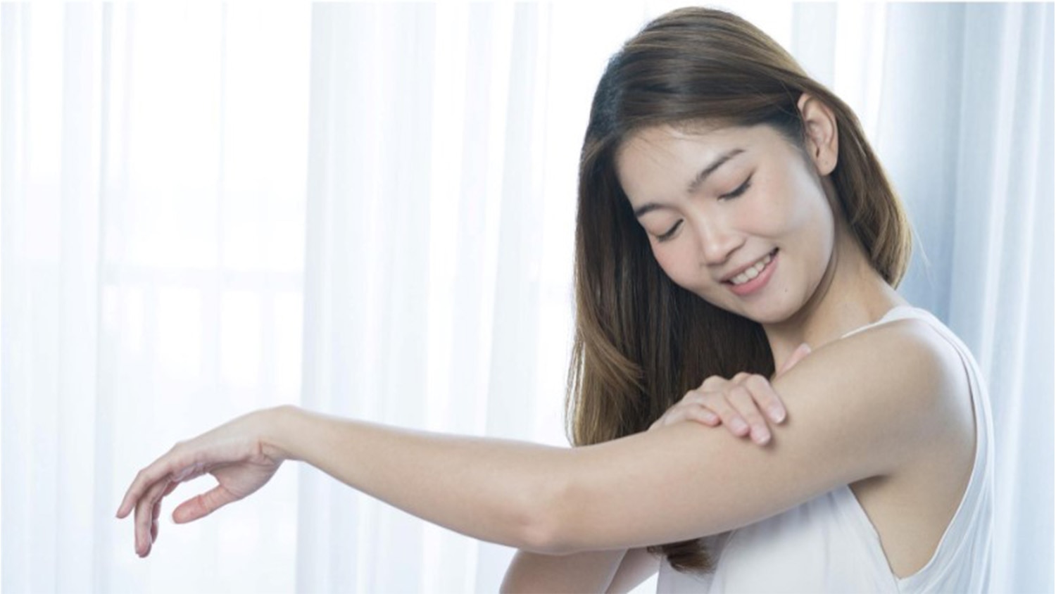  หญิงสาวกำลังทาครีมดูแลผิว ยิ้มอย่างอ่อนโยนขณะใช้มือสัมผัสแขนของเธอ