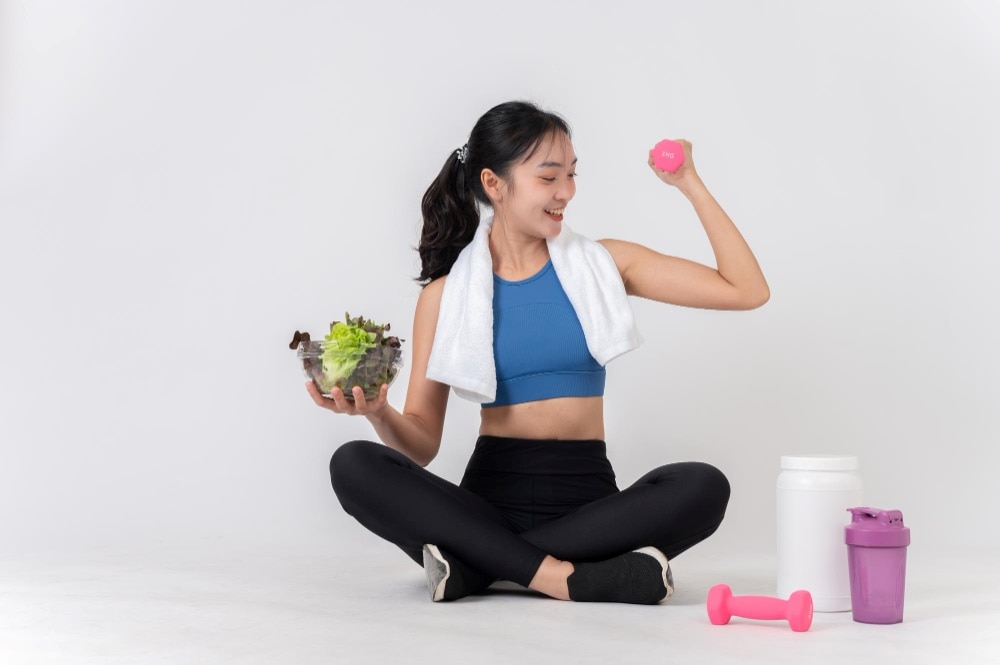 หญิงที่กำลังนั่งขัดสมาธิสวมเสื้อกีฬา ในมือซ้ายถือชามสลัดและในมือขวาถือลูกเหล็กสีชมพูขนาดเล็ก 